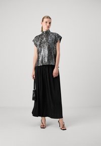Silkefarget sequinsbluse med høy krage, sammen med et svart plisserte maxiskjørt. Modellen holder en liten svart veske og har på seg høyhælte sandaler med stropper.