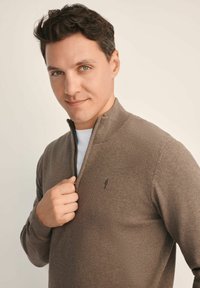 Brązowy sweter zapinany na zamek z żeberkowym kołnierzem, miękka tekstura i małe haftowane logo na klatce piersiowej, noszony na białej koszuli.
