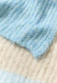 Puha kék kötött anyag világoszöld díszítéssel, bordázott mintázattal, krémszínű, bolyhos háttéren nyugszik.