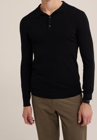 Svart långärmad pikétröja i stickad kvalitet, med klassisk krage, tvåknappssluiting och ribbade ärmslut, ihop med khakibyxor.