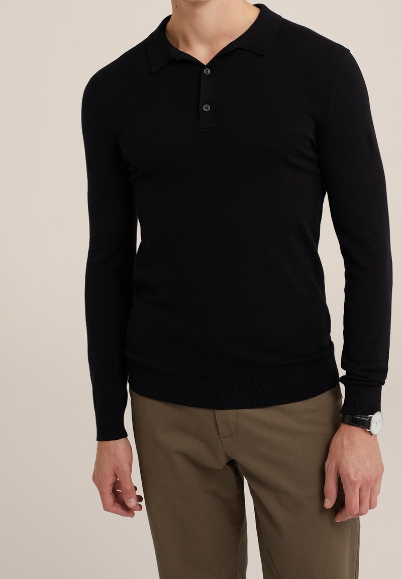 Svart långärmad pikétröja i stickad kvalitet, med klassisk krage, tvåknappssluiting och ribbade ärmslut, ihop med khakibyxor.