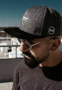 Graue Snapback-Basecap mit einem schwarzen Schirm, ausgestattet mit einem Diamond-Logo-Patch und einem weißen Markenemblem. Eine Sonnenbrille mit Metallrahmen rundet den Look ab.