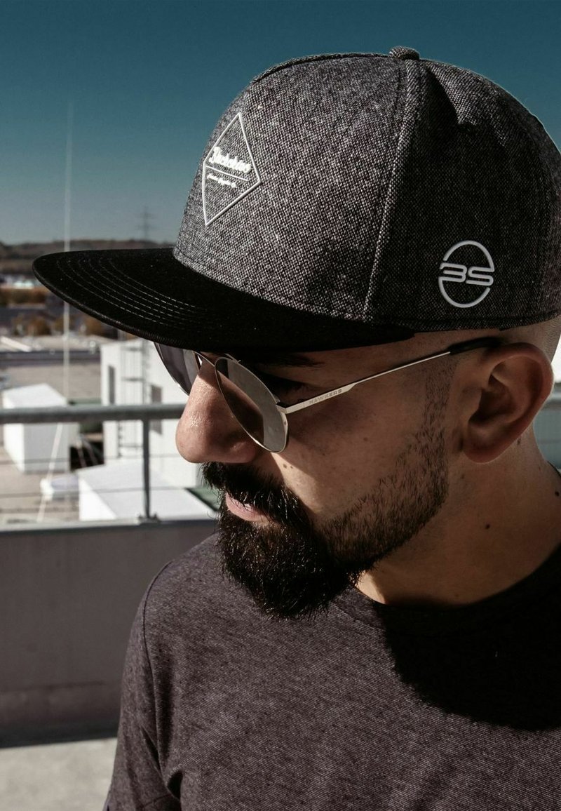 Siva snapback kapa s crnim vizirjem, koja ima zakrpu s dijamantnim logom i bijelim emblemom marke. Sunčane naočale s metalnim okvirom zaokružuju izgled.
