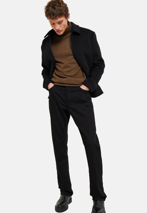 Veste noire avec col sur un pull en col roulé marron, associée à un pantalon noir et des bottes noires. Coupe ajustée, design minimaliste, texture lisse.