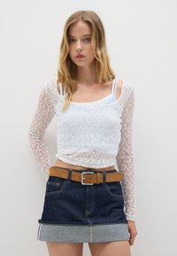 Top lungo bianco in pizzo con maniche, indossato sopra una canotta bianca, abbinato a una mini gonna di jeans con orlo azzurro chiaro e cintura marrone.