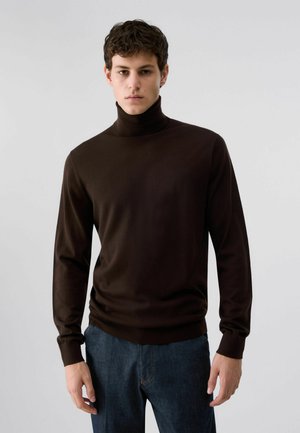 Giovane uomo che indossa un maglione dolcevita marrone scuro e jeans blu, in piedi davanti a uno sfondo chiaro e uniforme.