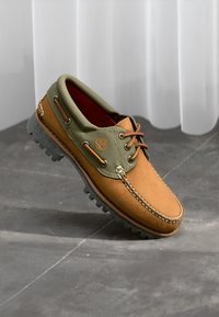 Braune Lederbootsschuhe mit olivgrünen Stoffakzenten, rotem Innenfutter und grauer Gummisohle. Verfügt über ein schnürbares Design und ein Logo-Emblem.