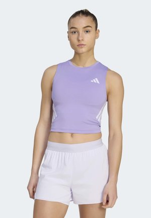 Jeune femme portant un crop top sportif lavande sans manches et un short blanc, debout avec une expression neutre devant un fond uni.