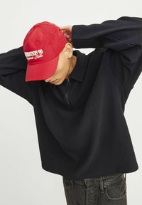 Pull noir à côtes avec col zippé un quart et coupe décontractée. Casquette rouge avec du texte blanc à l'avant. Jean foncé complètent la tenue.