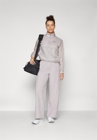 Light gray windbreaker with a half-zip and elastic waist, paired with light purple wide-leg trousers and a black duffel bag. White sneakers.