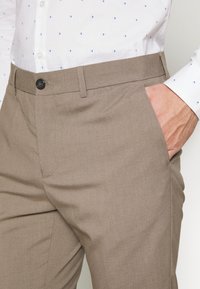 Light brown trousers with a smooth texture, featuring belt loops and a single front pocket, paired with a white shirt with blue patterns.