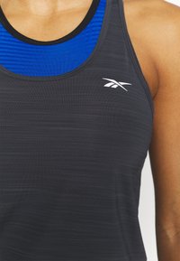 Czarny sportowy top bez rękawów z niebieską prążkowaną warstwą wewnętrzną, z białym logo Reebok na klatce piersiowej. Materiał ma subtelną teksturę.