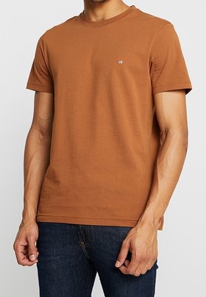 Mężczyzna w prostym brązowym t-shircie z krótkim rękawem z małym logo "ck" na piersi i ciemnoniebieskich dżinsach, stojący na jasnym tle.