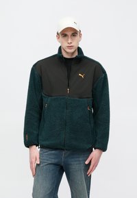 Vihreä fleecetakki, jossa musta paneeli, oransseja yksityiskohtia, vetoketjusuljenta ja kaksi sivutaskua. Päällä valkoinen lakki. Siniset farkut.