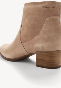 Bottine beige en suède avec talon bloc en bois et coutures apparentes, présentant un logo "tamaris" embossé près du bord supérieur.