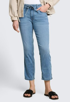 Kobieta w jasnoniebieskich krótkich jeansach z paskiem do wiązania, beżowej kurtce, białym topie i czarnych sandałach na płaskiej podeszwie, stojąca na jednolitym tle.