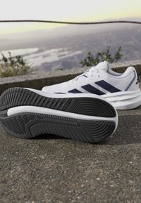 Chaussures de sport blanches avec semelles noires et rayures foncées reposant sur une surface en béton avec un fond extérieur flou.