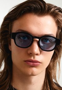 Black round sunglasses with opaque lenses and a thick frame, featuring a matte finish and subtle angular design. User's face partially visible.
