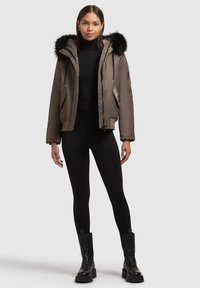 Braune Pufferjacke mit einer Kunstfellkapuze, Seitentaschen und elastischen Bündchen, kombiniert mit schwarzen Leggings und klobigen schwarzen Stiefeln.
