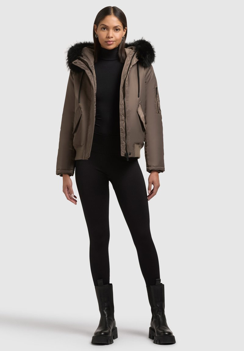Braune Pufferjacke mit einer Kunstfellkapuze, Seitentaschen und elastischen Bündchen, kombiniert mit schwarzen Leggings und klobigen schwarzen Stiefeln.