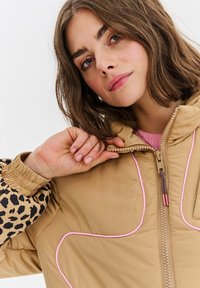 Žena s hnedými vlasmi po pleciach, oblečená v béžovej bunde s ružovým lemovaním a rukávmi s leopardím vzorom, drží golier bundy blízko svojho krku.