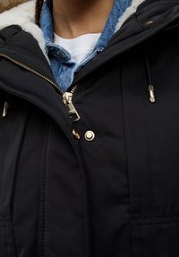 Giacca nera con zip, con accessori in oro, foderata in pelliccia sintetica e con texture trapuntata. Indossata sopra una camicia di jeans e una T-shirt bianca.