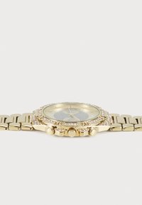 Montre dorée avec un cadran texturé et des accents en cristal. Elle possède un bracelet en métal et trois boutons sur le côté, affichant une forme ronde.