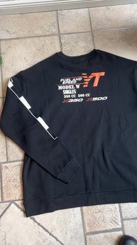 Fekete, hosszú ujjú póló fehér és narancssárga felirattal, amelyen a következők találhatók: "FUEL AND SPEED", "MODEL W SINGLES" és "350 CC 500 CC" a hátsó részen, fehér csíkkal az ujjakon.