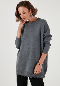Szary dzianinowy sweter o luźnym kroju, z ściągaczami przy mankietach i frędzlami u dołu. Miękka tekstura o minimalistycznym wyglądzie, idealny do warstwowego ubierania się.