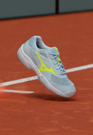 Világoskék és neon sárga sportcipő a levegőben, egy agyagos teniszpálya felett, melyen szétszórtan vörös agyagrészecskék és fehér pályavonalak láthatók.