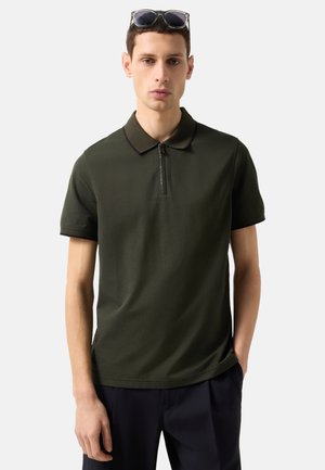 Mladý muž v tmavě zelené polokošili s krátkými rukávy a předním zipem, se slunečními brýlemi opřenými na hlavě, s rukama v kapse námořnických kalhot.