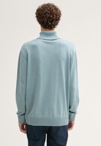 Persoon met krullend haar, gekleed in een lichtblauwe coltrui en donkere jeans, vanuit de achterkant bewonderd tegen een eenvoudige achtergrond.