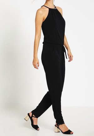 Mujer con un mono negro sin mangas con cintura atada y tacones abiertos negros con tacones metálicos tipo bloque.