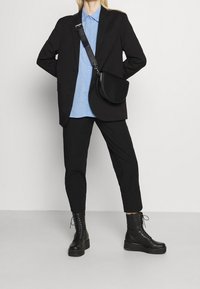 Personne portant un blazer noir, un pantalon noir, des bottes à plateforme noires à lacets, une chemise à boutons bleu clair et un sac bandoulière noir, le tout sur fond uni.