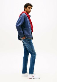Chaqueta de denim en azul oscuro con dos bolsillos frontales, superpuesta a una camisa de rayas en blanco y negro. Jeans azules ajustados y zapatillas blancas con detalles en rojo.