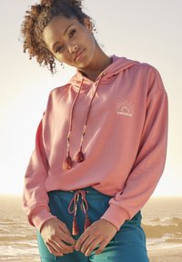 Felpa con cappuccio rosa dotata di cordini a nappina e stampa frontale che recita "SUNRISE PARADISE." Abbinata a pantaloni teal con cordini.