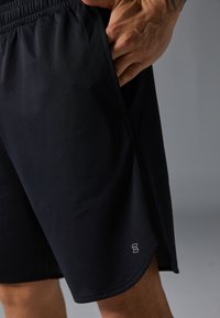 Svarte atletiske shorts med lett, pustende stoff. Tilgang til sidelommer og en liten logo nær kanten. Teksturert meshdesign.
