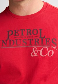 Červené bavlněné tričko s výrazným černým grafickým potiskem, na kterém je nápis "PETROL INDUSTRIES & Co" spolu s "registrováno od 1989".