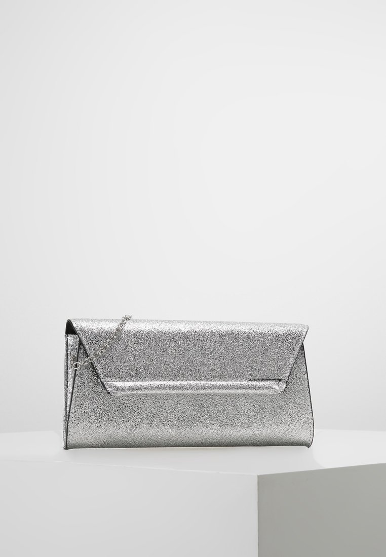 Pochette pailletée argentée avec rabat triangulaire et fine chaîne, posée sur une surface blanche sur un fond uni.