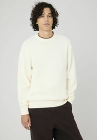 Pull en crème avec un tricot texturé, encolure ronde et poignets côtelés. Associé à un pantalon marron foncé, mettant en avant une coupe décontractée.