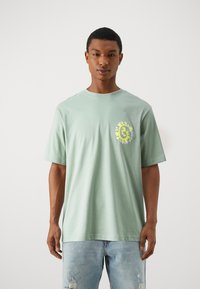 Camisa de algodón verde claro con mangas cortas, con un escote redondo y un gráfico circular en amarillo y blanco en el área izquierda del pecho.
