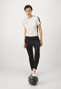 Biela tričko Nike s čiernymi akcentmi, skombinované s čiernymi nohavicami, ktoré majú fialové logo a emblém. Model stojí na futbalovej lopte.