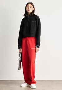 Czarna rozpinana bluza z logo, w parze z luźnymi czerwonymi spodniami dresowymi z logo. Modelka trzyma srebrną torebkę i ma na sobie białe trampki.