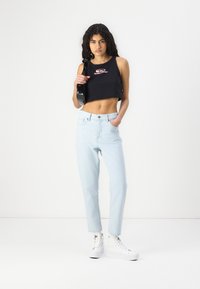 Young woman standing against white background wearing black crop top, light blue jeans, white sneakers, and holding black handbag on shoulder.