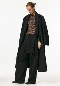 Palton negru din lână cu nasturi mari, purtat peste un top transparent din dantelă și o fustă asimetrică din dantelă, asortate cu pantaloni largi negri și pantofi comozi.