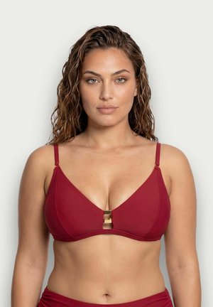 Mujer con cabello ondulado que lleva un top de bikini rojo con detalles dorados, mirando hacia adelante sobre un fondo claro y liso.