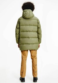 Person med kort, lockigt hår som bär en grön, huvad puffjacka, ljusbruna mönstrade byxor och svarta sneakers, stående med ryggen mot kameran.