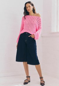 Maglione a maglia aperta di colore rosa brillante con maniche lunghe, abbinato a culottes in denim scuro. Le calzature presentano un dettaglio con cinturino e fibbia.