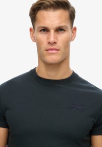 Sort bomulds t-shirt med korte ærmer, rund hals og broderet logo i lilla på venstre brystområde. Glat tekstur.