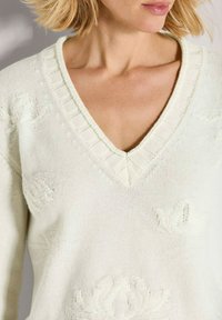Vrouw in een crèmekleurige V-hals gebreide trui met textuurdecoraties van bloemen en kort blond haar, afgebeeld van de nek tot halverwege de torso.